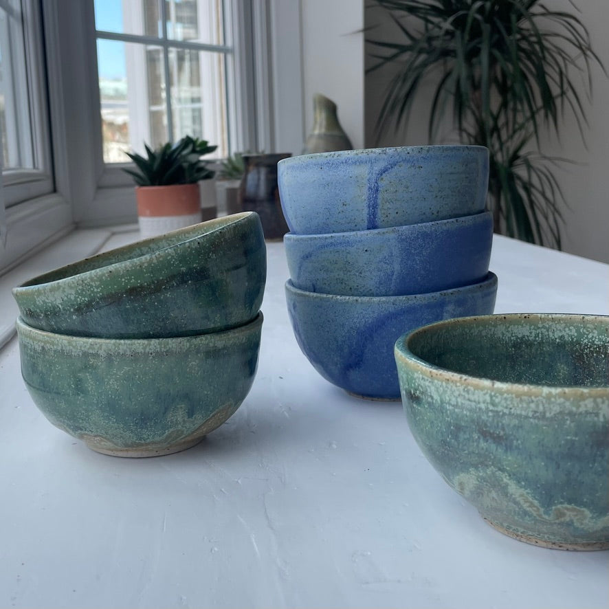 A set of handmade ceramic bowls with a rustic glaze, including two stacked bowls in green, a stack of three in blue, and one single bowl in the foreground, displayed on a white table near a window with plants in the background.