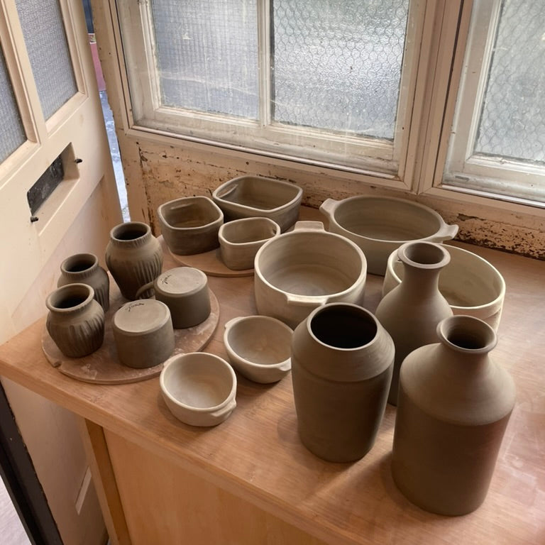 A collection of handmade clay pottery vessels, including vases, pots, and bowls, arranged on a wooden table in front of a window.