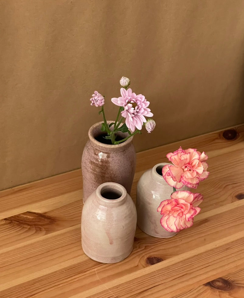 Three small ceramic vases in neutral earthy tones arranged on a wooden surface against a tan backdrop, with pink flowers placed in two of the vases.