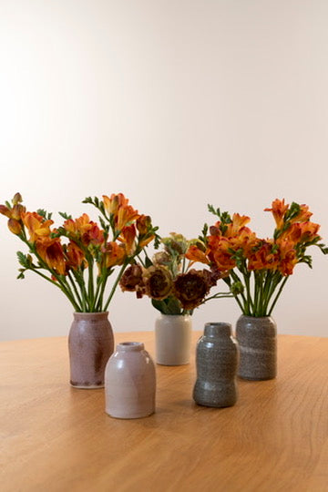 Five small ceramic vases in muted earthy tones arranged on a light wood table, with orange and brown flowers placed in two of the vases against a plain off-white wall.