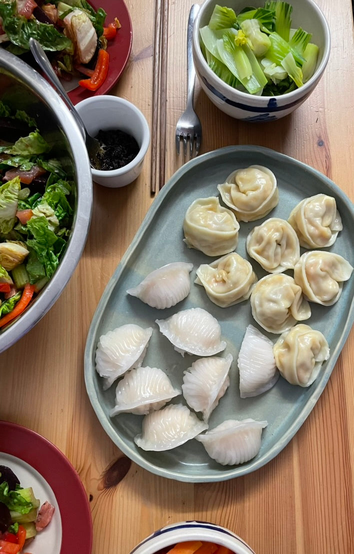 A top-down view of a wooden dining table featuring a light blue-gray oval platter filled with dumplings, surrounded by various colorful salads in bowls and plates.