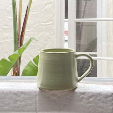 A light sage green ceramic mug sits on a white tiled windowsill next to a large green banana leaf plant, with a textured white wall and window frame in the background.