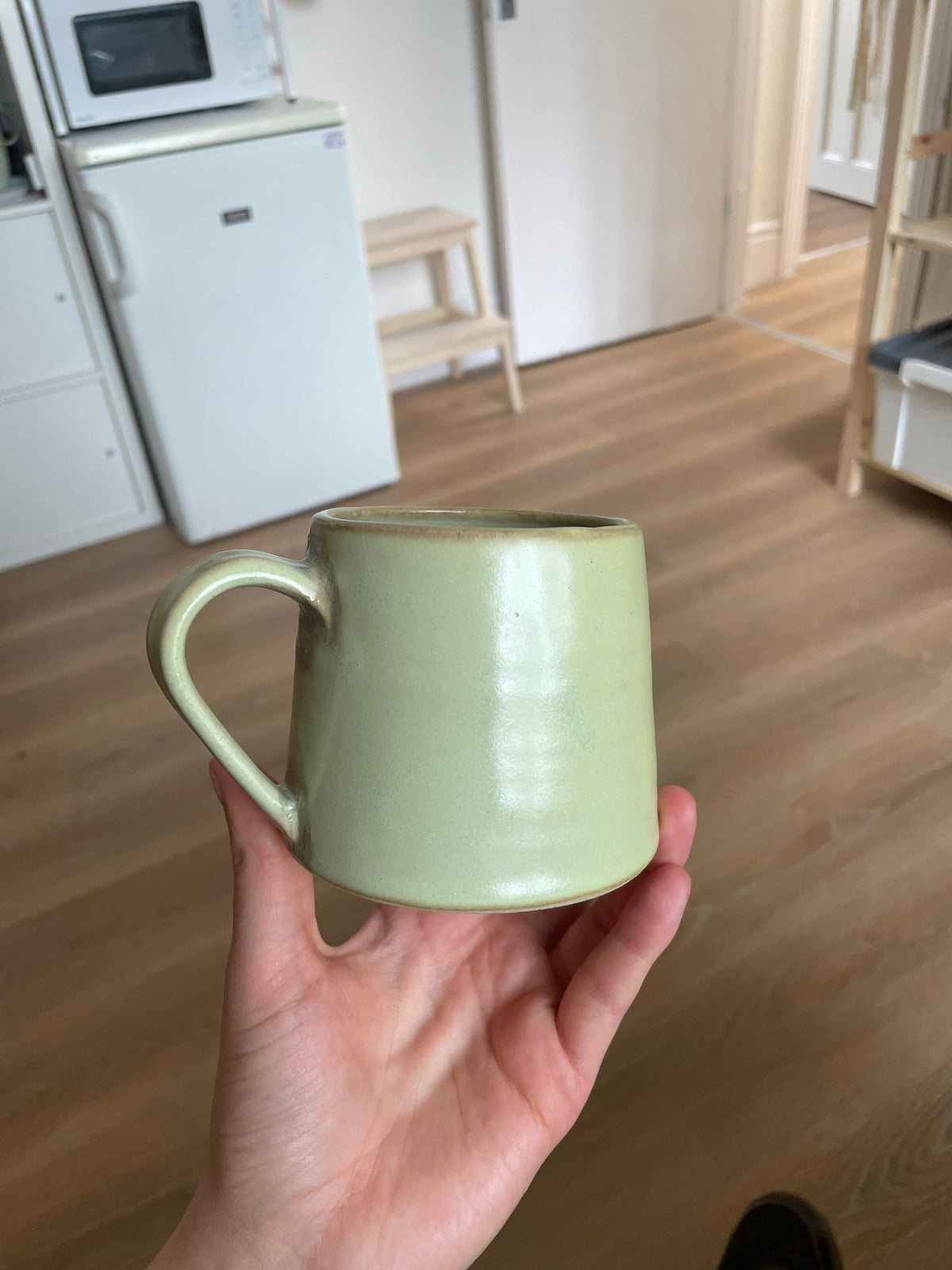 A light green ceramic mug with a rounded cylindrical shape and a loop handle, held by a person's hand in a kitchen setting with wooden floors and white appliances in the background.