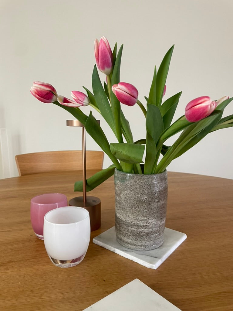 A bouquet of pink tulips in a textured gray ceramic vase, styled on a wooden dining table alongside two small candles and a white coaster.