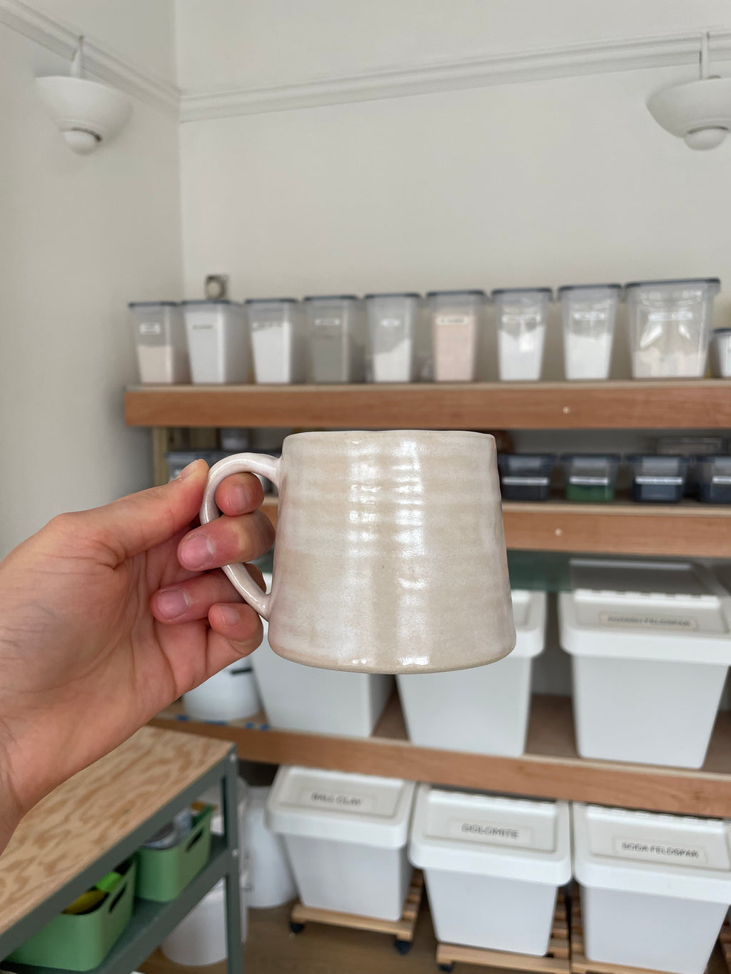 A hand holding a handmade ceramic mug with a textured, ribbed surface in a warm off-white or cream color, set against a background of shelves containing white storage containers.
