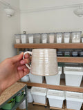 A hand holding a handmade ceramic mug with a textured, ribbed surface in a warm off-white or cream color, set against a background of shelves containing white storage containers.