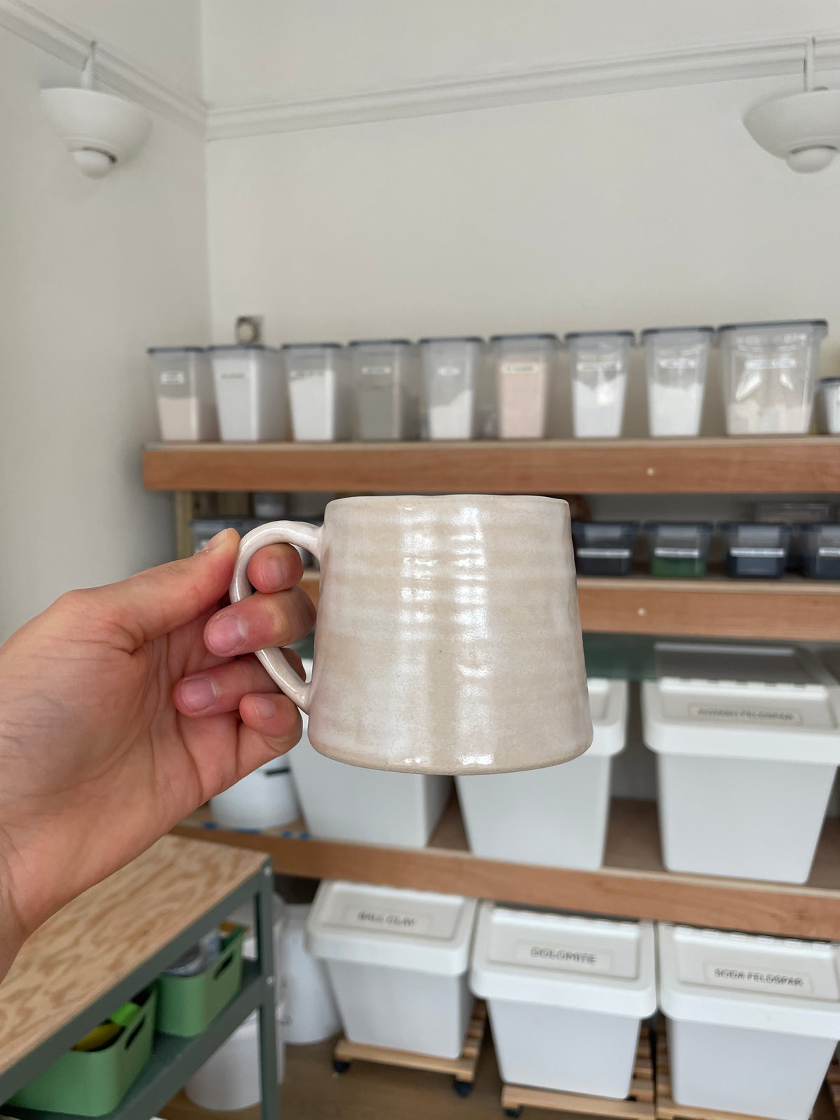 A hand holding a handmade ceramic mug with a textured, ribbed surface in a warm off-white or cream color, set against a background of shelves containing white storage containers.